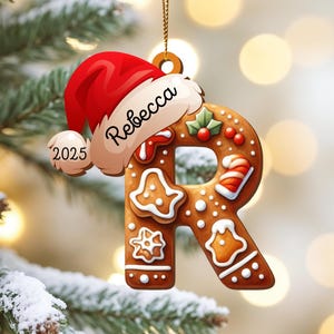 May include: A gingerbread-style Christmas ornament shaped like the letter "R", decorated with icing and festive candies. It is topped with a Santa hat that has the name "Rebecca" and the year "2025" on it. The ornament hangs from a gold string.