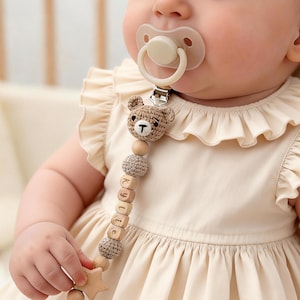 Puede incluir: Un chupete para bebé con tetina beige y clip. El clip está sujeto a un sujetachupetes de madera y ganchillo con cabeza de oso y el nombre "Kylie" escrito con bloques de madera. Una estrella de madera en el extremo.