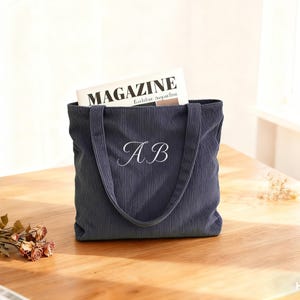 May include: Navy blue corduroy tote bag with white embroidered initials "AB". The bag has a rectangular shape with two handles and is filled with a magazine. The word "MAGAZINE" is visible on the magazine.