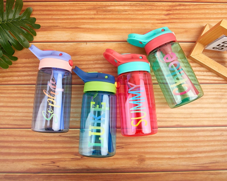 May include: Four colorful reusable water bottles with personalized names on them. The bottles are blue, green, red, and turquoise. The bottles are on a wooden surface.