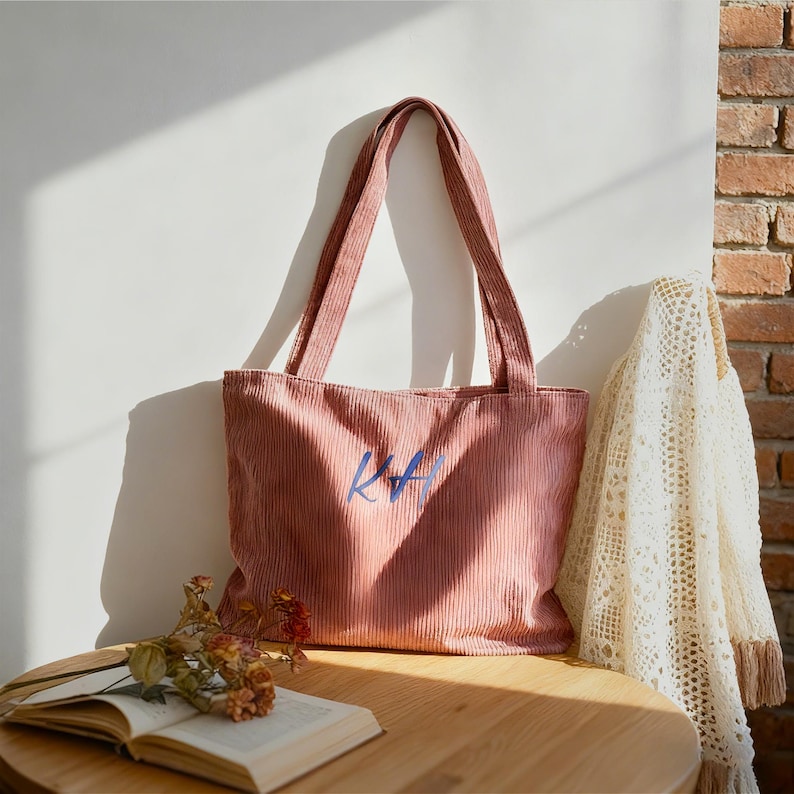 May include: A dusty rose corduroy tote bag with long handles. The bag has the blue letters "KA" embroidered on the front. The bag is sitting on a wooden table with a book and flowers. A white lace shawl hangs in the background.