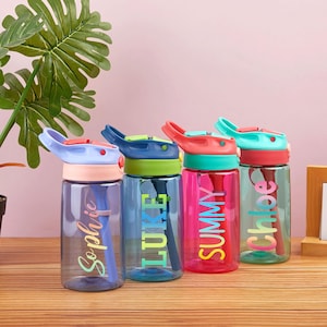 May include: Four colorful reusable water bottles with flip top lids and built-in straws. The bottles are labeled with the names "Sophia", "Luke", "Summy", and "Chloe" in a rainbow font.