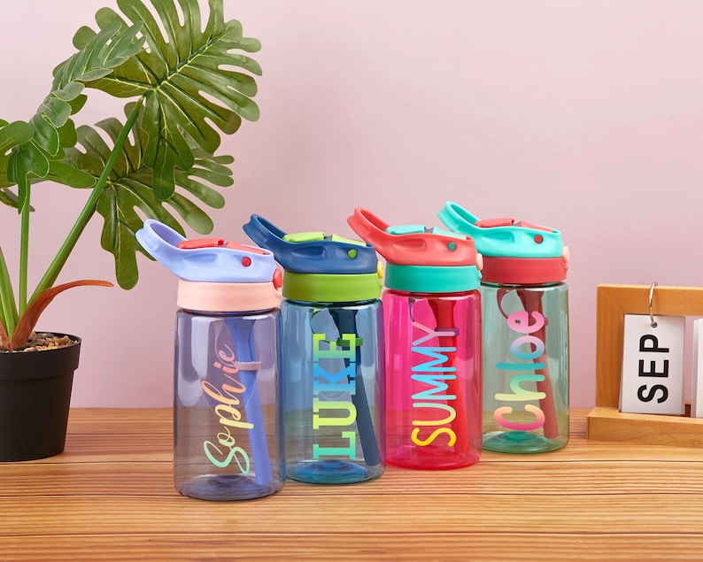May include: Four colorful reusable water bottles with flip top lids and built-in straws. The bottles are labeled with the names "Sophia", "Luke", "Summy", and "Chloe" in a rainbow font.