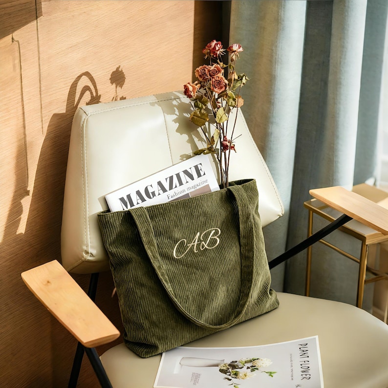 May include: Olive green corduroy tote bag with embroidered "AB" initials. The bag is filled with a magazine and dried flowers, resting on a chair. The word "MAGAZINE" is visible on the magazine.