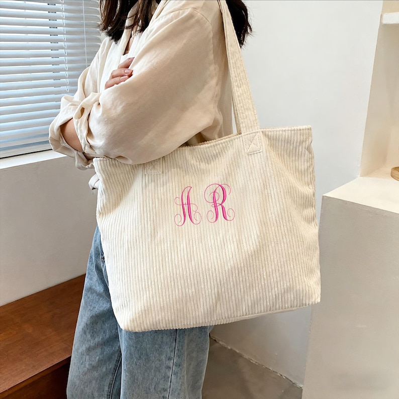 May include: Cream-colored corduroy tote bag with pink embroidered initials "AR". The bag has a rectangular shape with two long handles. The corduroy fabric has vertical ridges. The bag is held by a person.
