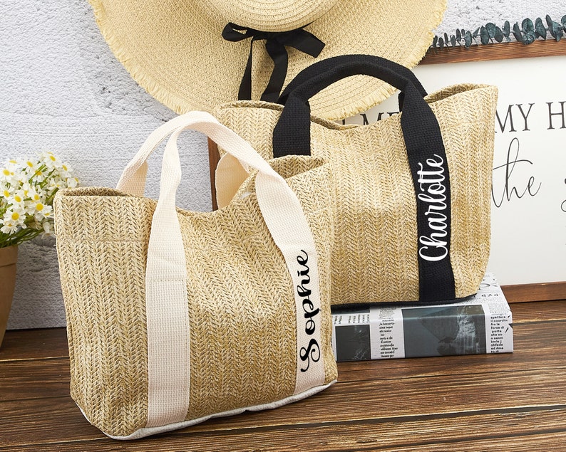 Puede incluir: Dos bolsas de mano de paja con nombres personalizados. La bolsa de la izquierda tiene el nombre "Sophie" bordado en la correa blanca. La bolsa de la derecha tiene el nombre "Charlotte" bordado en la correa negra.
