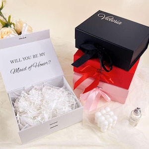 May include: White gift box with the text "WILL YOU BE MY Maid of Honor?" and shredded paper. Stacked gift boxes in black, red, and pink with ribbons. The black box has the name "Victoria" on the lid.