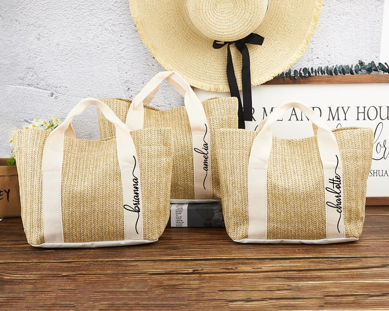 Puede incluir: Tres bolsas de mano de paja con correas blancas y nombres en escritura negra: Brianna, Amelia y Charlotte.