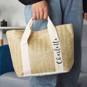 K&ouml;nnte beinhalten: Eine beige Tote Bag mit wei&szlig;en Henkeln und dem Namen "Charlotte" auf der Seite.