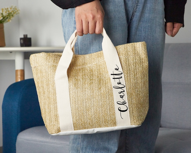 K&ouml;nnte beinhalten: Eine beige Tote Bag mit wei&szlig;en Henkeln und dem Namen "Charlotte" auf der Seite.