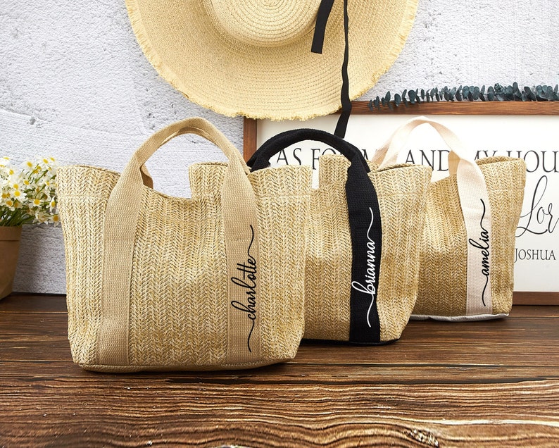 Puede incluir: Tres bolsas de mano de paja con nombres personalizados. Las bolsas son de color beige con letras negras y blancas. Los nombres son "Charlotte", "Brianna" y "Amelia".