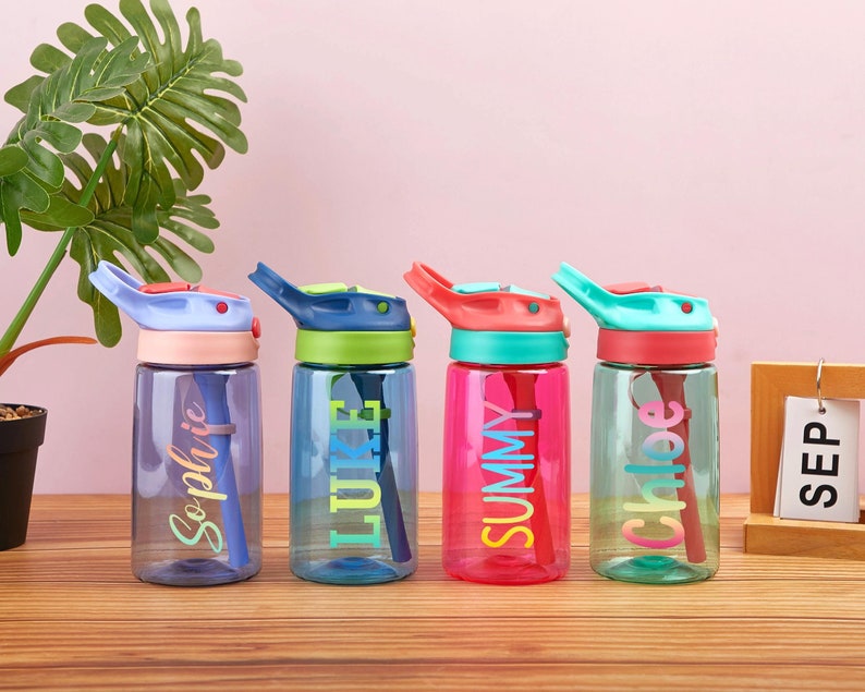 May include: Four colorful reusable water bottles with flip top lids and built-in straws. The bottles are personalized with the names "Sophie", "Luke", "Summy", and "Chloe" in a rainbow font.
