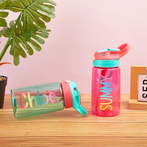 May include: Two colorful water bottles with personalized names. The bottle on the left is green with the name "Chloe" in rainbow letters. The bottle on the right is pink with the word "Summer" in rainbow letters.