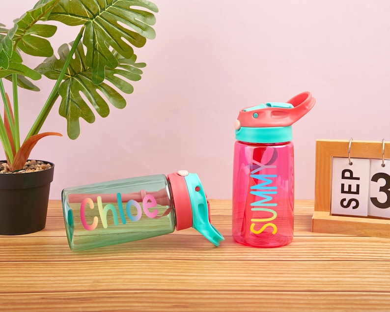 May include: Two colorful water bottles with personalized names. The bottle on the left is green with the name "Chloe" in rainbow letters. The bottle on the right is pink with the word "Summer" in rainbow letters.