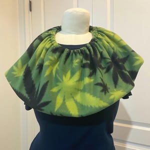 May include: A green and black fleece cowl neck warmer with a cannabis leaf pattern. The neck warmer is draped over a mannequin, showcasing its design and shape. The background is a plain white door.