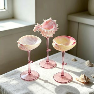 May include: Three stemmed glasses featuring bowls shaped like seashells in shades of pink, cream, and white. Each glass has a slender, translucent pink stem and a circular base. The central glass has a spiky, textured shell rim.