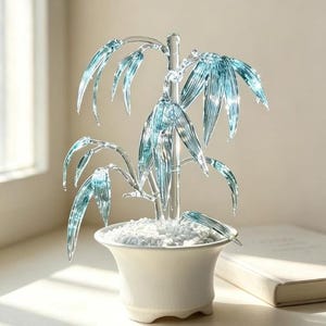 May include: A decorative glass sculpture of a bamboo plant with translucent light blue leaves and a clear stem. The plant is set in a white ceramic pot filled with small white stones, creating a clear and bright display of glass artistry.