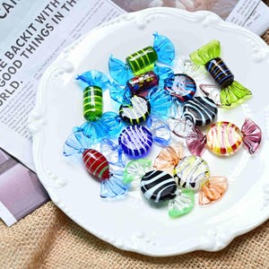 Pode incluir: Uma coleção de pequenos ornamentos de vidro em forma de rebuçado sobre um prato branco com bordas recortadas. As peças de vidro apresentam vários padrões às riscas e sólidos em tons de azul, verde, vermelho, amarelo, preto e transparente.
