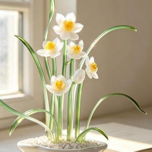 May include: A decorative glass sculpture of a cluster of white and yellow daffodils with translucent green stems and leaves, planted in a white ceramic pot filled with small white stones, creating a realistic floral arrangement display.