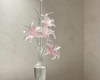 May include: A clear glass vase containing five delicate, light pink glass lily flowers with slender green stems. The flowers feature translucent, curved petals and pointed tips. A single detached pink glass lily flower rests nearby.