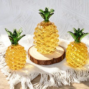 May include: Three decorative glass pineapple figurines featuring translucent yellow bodies with a textured diamond pattern and solid green glass leaves at the top. The central pineapple is elevated on a circular wooden slice base.