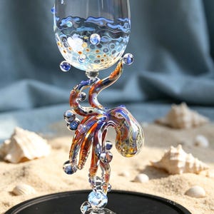 May include: A clear glass goblet featuring a stem shaped like a multi-colored octopus with orange, blue, and purple hues. The bowl of the glass is decorated with blue wave patterns and small, raised, multi-colored glass bubbles near the base.