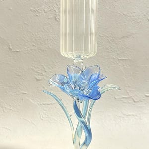May include: A clear glass candle holder featuring a vertical ribbed cylindrical top section supported by a stem shaped like a blue and light blue glass flower with curved petals and leaves, all resting on a circular clear glass base.