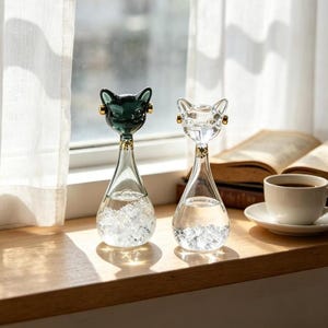 May include: Two glass cat-shaped bottles with gold-toned accents on the ears and neck. One bottle is made of dark green glass, and the other is clear glass. Both bottles have a teardrop-shaped base filled with small, clear, faceted glass beads.