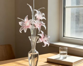 May include: A clear glass vase containing five delicate, light pink glass lily flowers with slender stems. The flowers feature translucent petals with subtle white tips. One additional detached pink glass lily flower rests on the surface nearby.