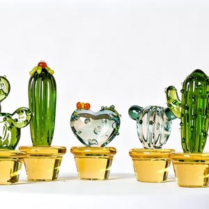 May include: A collection of five glass cactus figurines, each planted in a translucent yellow pot. The cacti vary in shape, including round, heart-shaped, and tall columnar forms, rendered in shades of green and clear glass with small raised details.