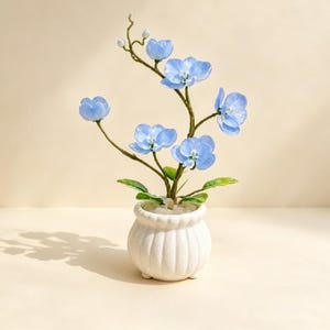 Puede incluir: Una planta de orquídea artificial con pétalos de color azul claro y tallos de color verde oscuro, que presenta varias flores abiertas y pequeños capullos. La planta está en una maceta de cerámica blanca, redonda, con textura acanalada y acabado moteado.