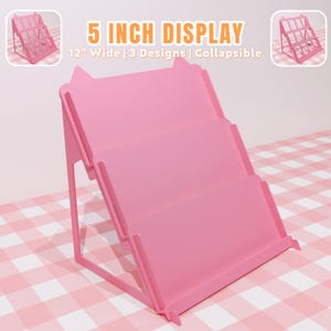 Peut inclure: Présentoir rose vif à trois niveaux. Le présentoir mesure 30.5 cm de large et est pliable. Le texte "5 INCH DISPLAY" est en haut. Le présentoir est sur une surface à carreaux roses et blancs.