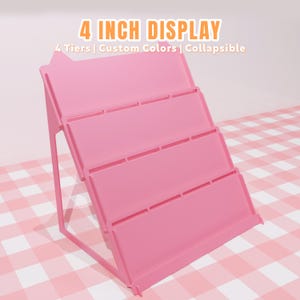 May include: A pink, four-tiered display stand with a collapsible design. The stand is 10.16 cm in height and features custom colour options. The text "4 INCH DISPLAY" is at the top, with additional text below.