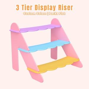 May include: A three-tiered display riser in pastel colours. The pink supports hold shelves in lavender, blue, and yellow. The text on the image reads "3 Tier Display Riser, Custom Colors | Packs Flat."