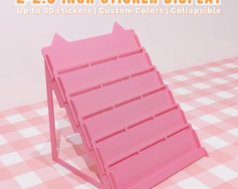 May include: A pink sticker display with multiple shelves. The display is designed to hold stickers that are 5.08-6.35 cm in size. The text on the display reads "2-2.5 INCH STICKER DISPLAY" and "Up to 30 stickers | Custom Colors | Collapsible".