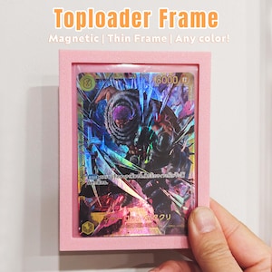 Peut inclure: Un cadre rose pour toploader contenant une carte de collection avec du texte japonais et une illustration colorée. Le cadre porte l'inscription "Toploader Frame" et "Magnetic | Thin Frame | Any color!"