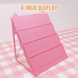 Puede incluir: Un expositor rosa de varios niveles con el texto "4 INCH DISPLAY FILES ONLY" en naranja. El expositor tiene cuatro estantes y un marco de soporte. El expositor está sobre una superficie a cuadros rosa y blanco.