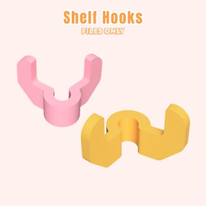 Pode incluir: Dois ganchos de prateleira, um rosa e outro amarelo, são exibidos contra um fundo de pêssego claro. Os ganchos têm formato de U com uma base circular. O texto "Shelf Hooks FILES ONLY" está em laranja na parte superior da imagem.