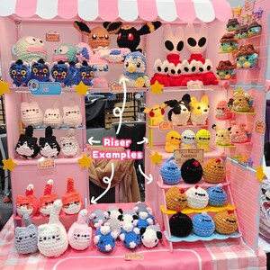 May include: A display of handmade crochet plush toys, including various characters and animals. The display features a pink and white booth with multiple shelves, showcasing the colorful creations. The toys include cats, penguins, and other creatures.