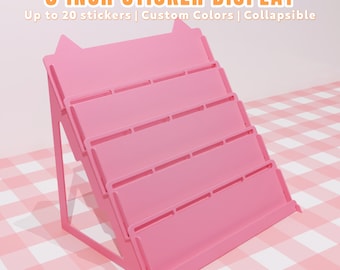 May include: A pink, collapsible sticker display with four angled shelves. The display is 3 inches wide and can hold up to 20 stickers. Text on the image reads "3 INCH STICKER DISPLAY" and "Up to 20 stickers | Custom Colors | Collapsible."