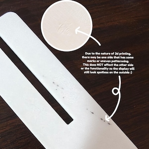 May include: Close-up of a white 3D-printed object with a rectangular shape and a cut-out section. The text in the image states that due to the 3D printing process, one side may have marks. The other side will look spotless.