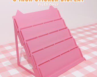 May include: A pink, tiered sticker display with the text "3 INCH STICKER DISPLAY FILES ONLY". The display has four shelves and is designed to hold stickers. The display is approximately 3 inches wide.