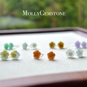 Puede incluir: Un conjunto de seis pares de pendientes de jade con forma de flor en varios colores, incluyendo verde, naranja y lavanda. Los pendientes se muestran sobre una superficie blanca con un marco de madera.