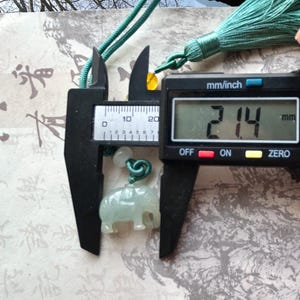 May include: A light green jade elephant pendant, 2.14 mm in size, is measured with a digital caliper. The pendant is suspended from a green cord with a tassel. The background features Asian calligraphy.
