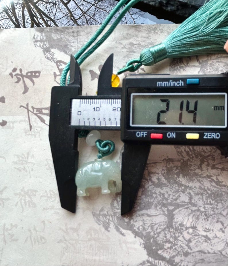 May include: A light green jade elephant pendant, 2.14 mm in size, is measured with a digital caliper. The pendant is suspended from a green cord with a tassel. The background features Asian calligraphy.