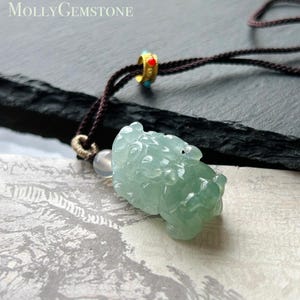 Könnte beinhalten: Ein hellgrüner Jade-Anhänger in Form eines Fabelwesens, an einer braunen Kordel mit einer goldfarbenen Perle befestigt. Der Anhänger ist aufwendig geschnitzt und hat eine durchscheinende Qualität. Der Text "MollyGemstone" ist oben links zu sehen.