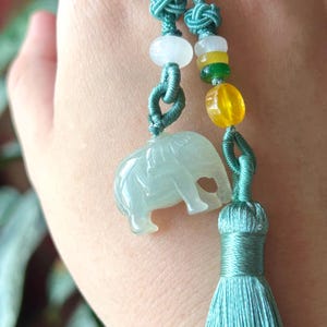 May include: A jade elephant pendant with a teal tassel and cord. The pendant is light green and has a detailed carving of an elephant. The cord features colorful beads and decorative knots. This item is a unique piece of jewelry.