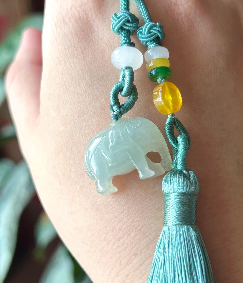 May include: A jade elephant pendant with a teal tassel and cord. The pendant is light green and has a detailed carving of an elephant. The cord features colorful beads and decorative knots. This item is a unique piece of jewelry.