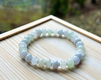May include: A beaded bracelet featuring a mix of pale green, white, and lavender-colored stones. The bracelet is strung on a flexible cord and has small silver-colored spacers between each bead. The bracelet is displayed on a light wooden surface.