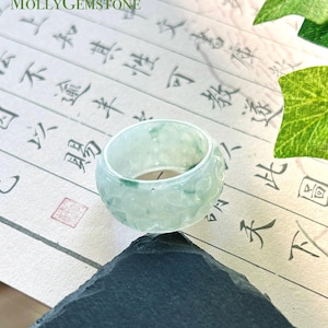 Puede incluir: Un anillo de jade verde pálido con un patrón floral tallado alrededor de la banda exterior. El anillo tiene un acabado liso y pulido con sutiles inclusiones de color verde oscuro en toda la piedra. El texto MOLLYGEMSTONE es visible arriba a la izquierda.